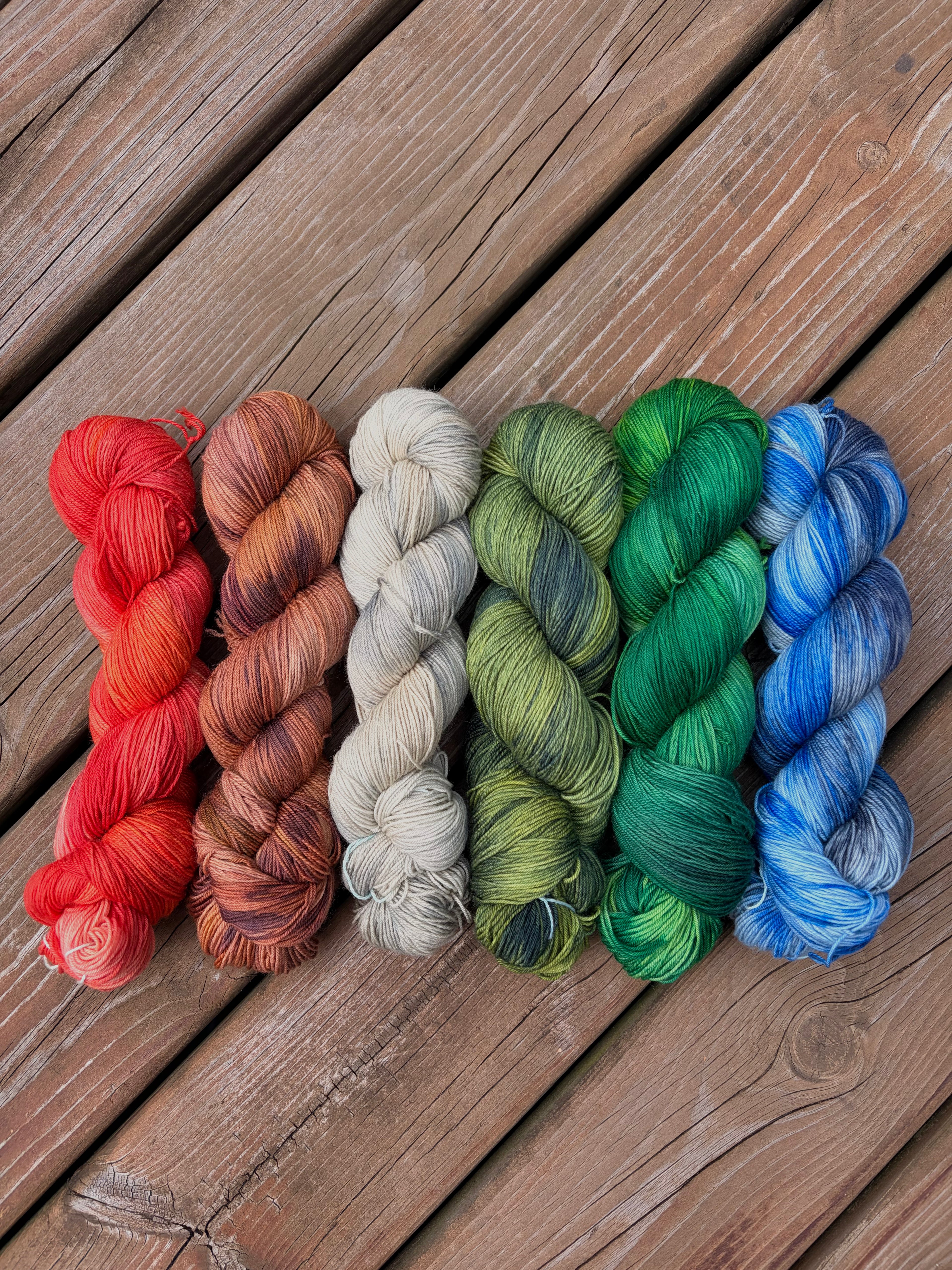 Six skeins of variegated yarn arranged side by side on a rustic wooden plank surface. The yarn colors include red-orange, terracotta, cream, olive green, emerald green, and blue with white streaks.
