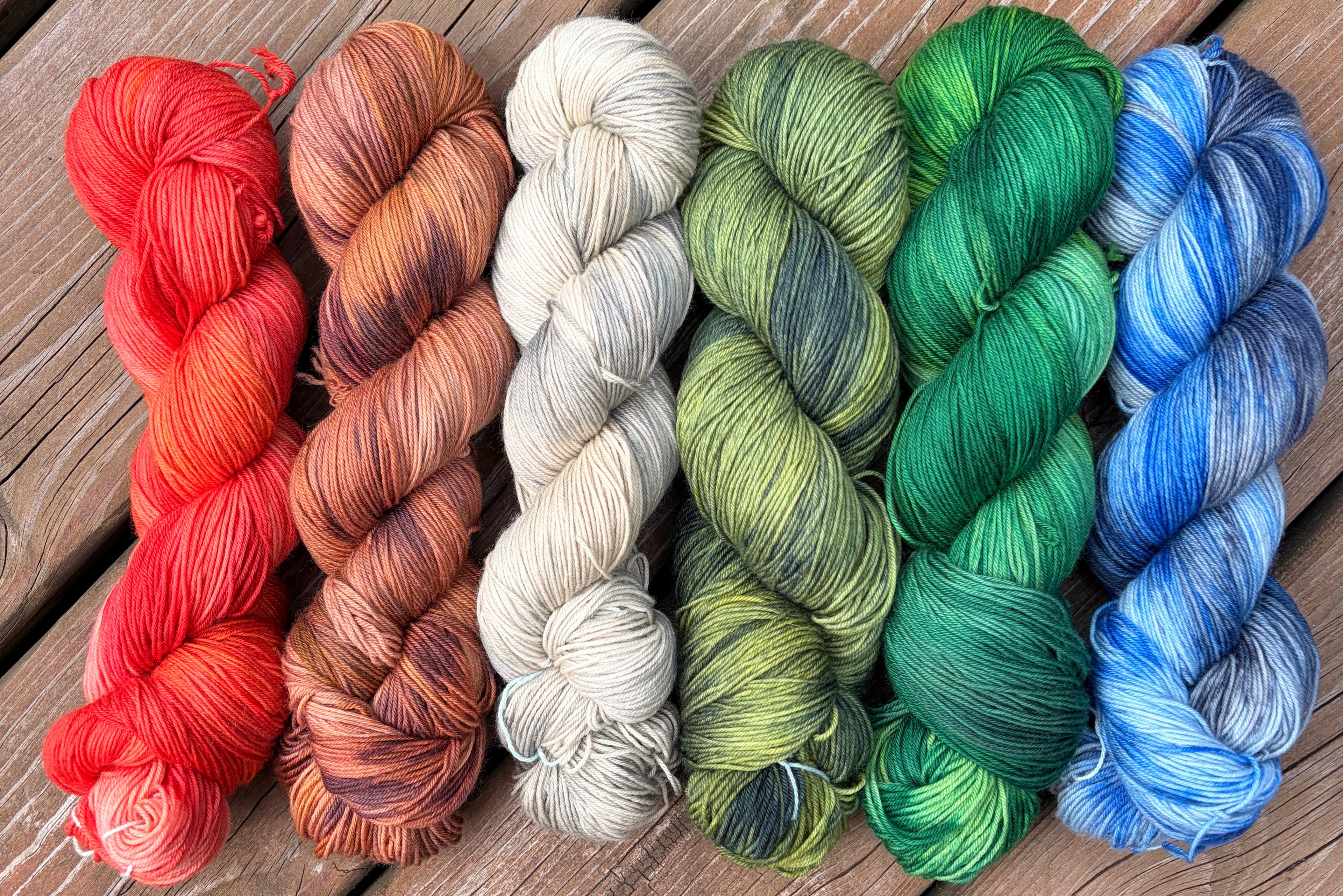 Six twisted skeins of variegated yarn arranged side by side on a rustic wooden plank surface. The yarn colors progress from bright red and orange, through copper and cream, to olive and emerald greens, and finish with blue and gray variegation.